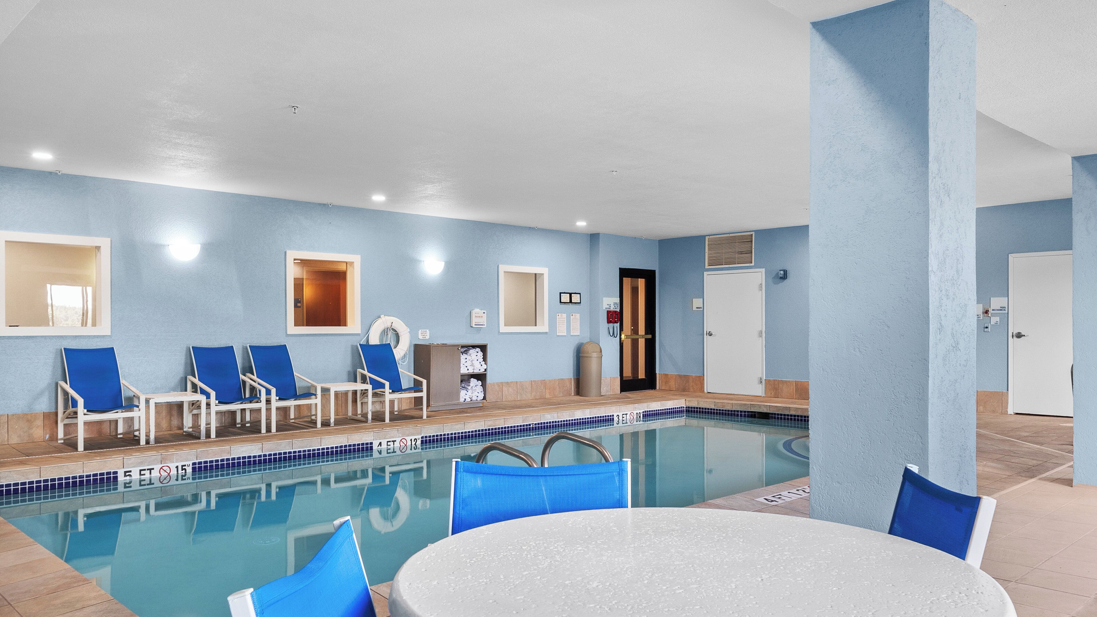 An indoor pool area with blue chairs, a white table, and a door leading to a changing room.