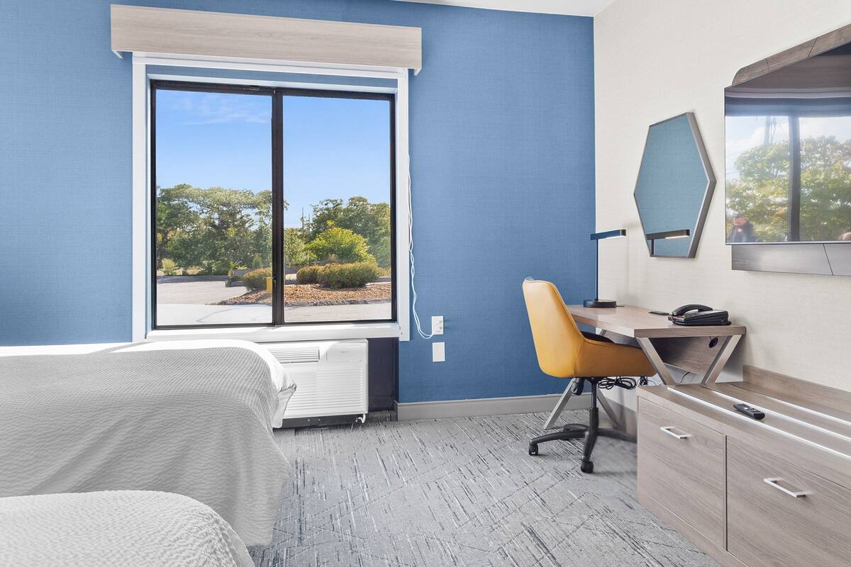 A hotel room with a bed, a desk with a chair, a large window, and a wardrobe.
