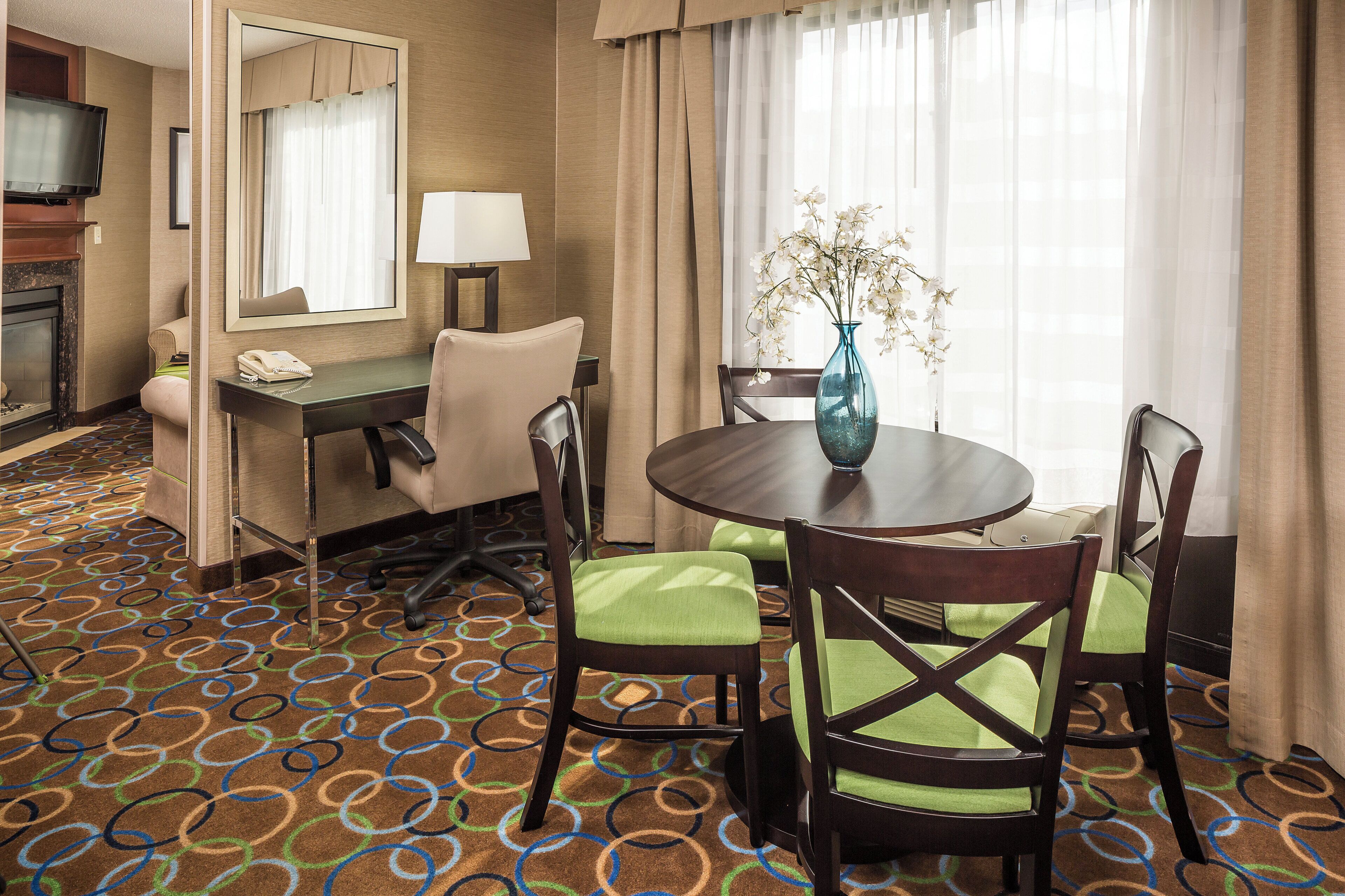 A hotel room with a dining area featuring a round table, four chairs, a vase with flowers, a desk with a lamp, a telephone, and a television on a stand.
