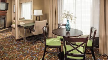 A hotel room with a dining area featuring a round table, four chairs, a vase with flowers, a desk with a lamp, a telephone, and a television on a stand.