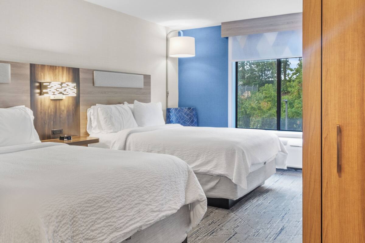 A hotel room with two beds, a nightstand, a lamp, and a window with a view of greenery.