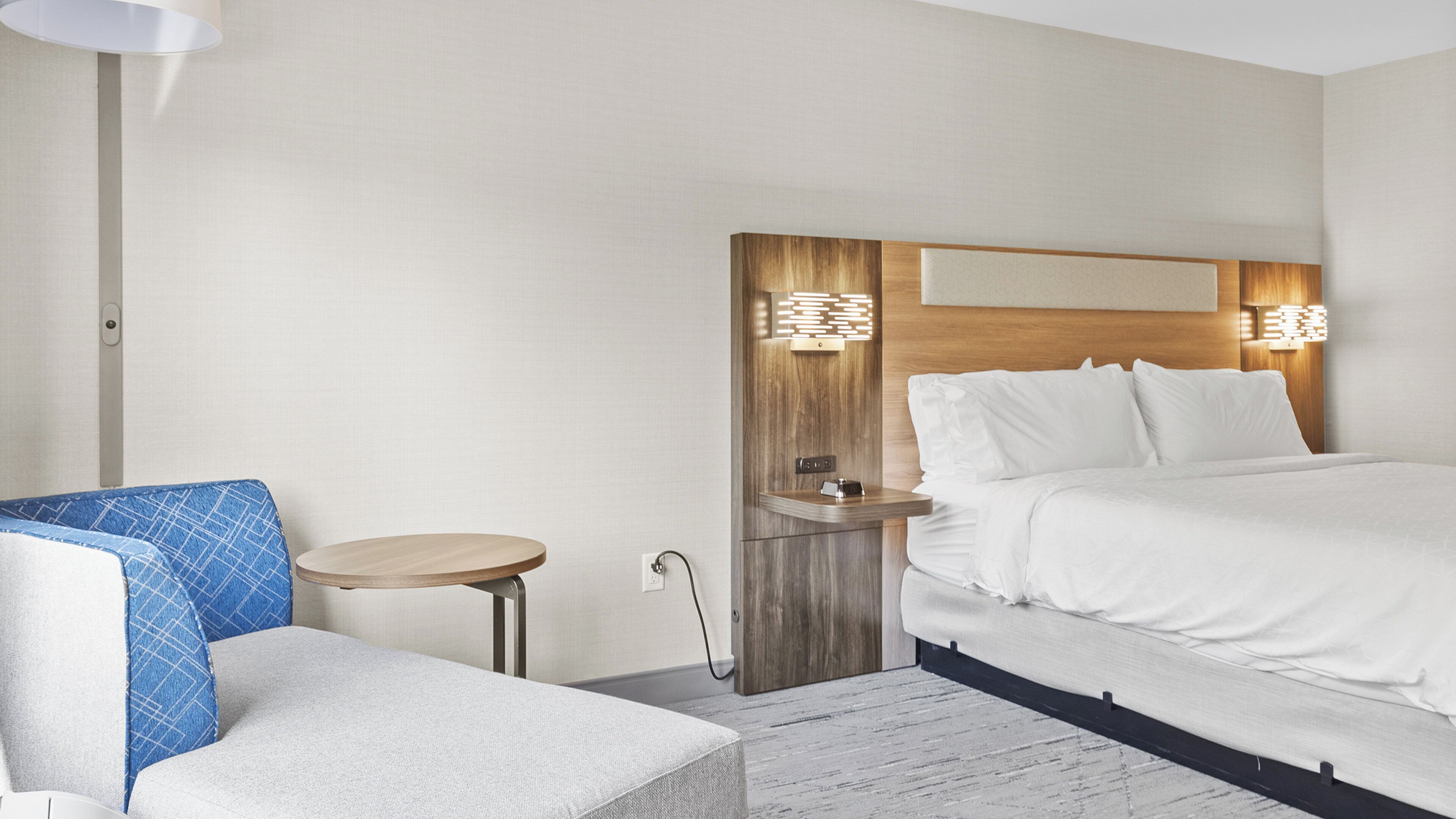 Une chambre d’hôtel avec un lit, une petite table ronde, une chaise et une lampe fixée au mur.