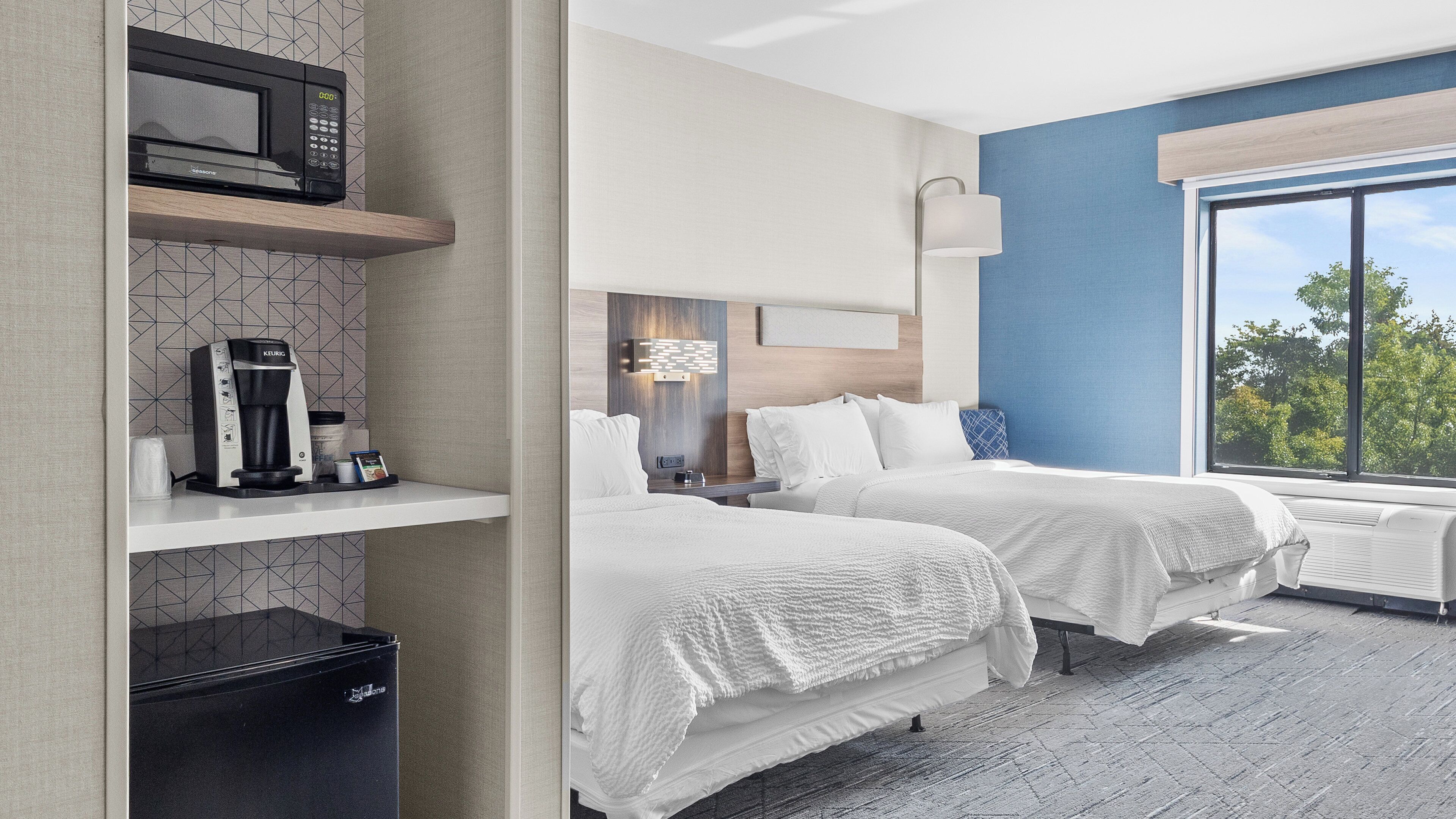 A hotel room with two beds, a coffee machine, a microwave, and a large window with a view of trees.