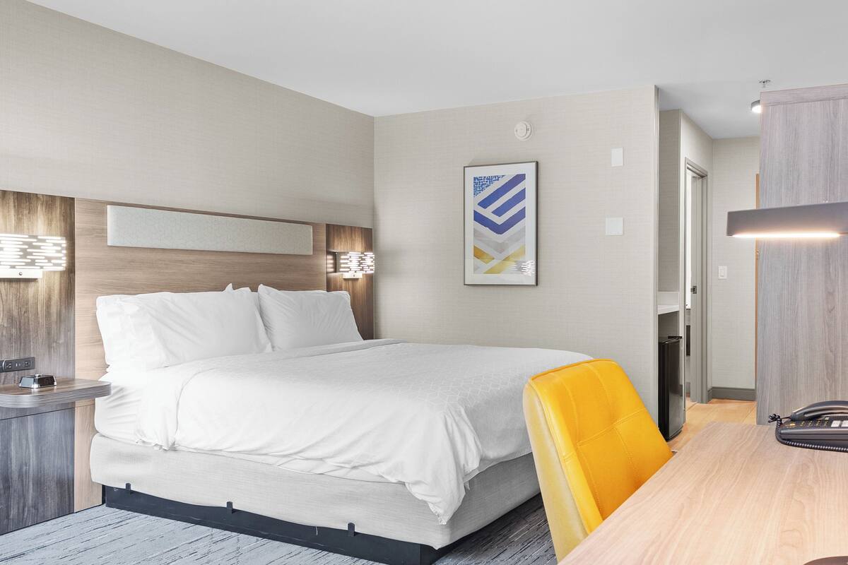 A modern hotel room with a bed, bedside table, lamp, and a desk with a chair.
