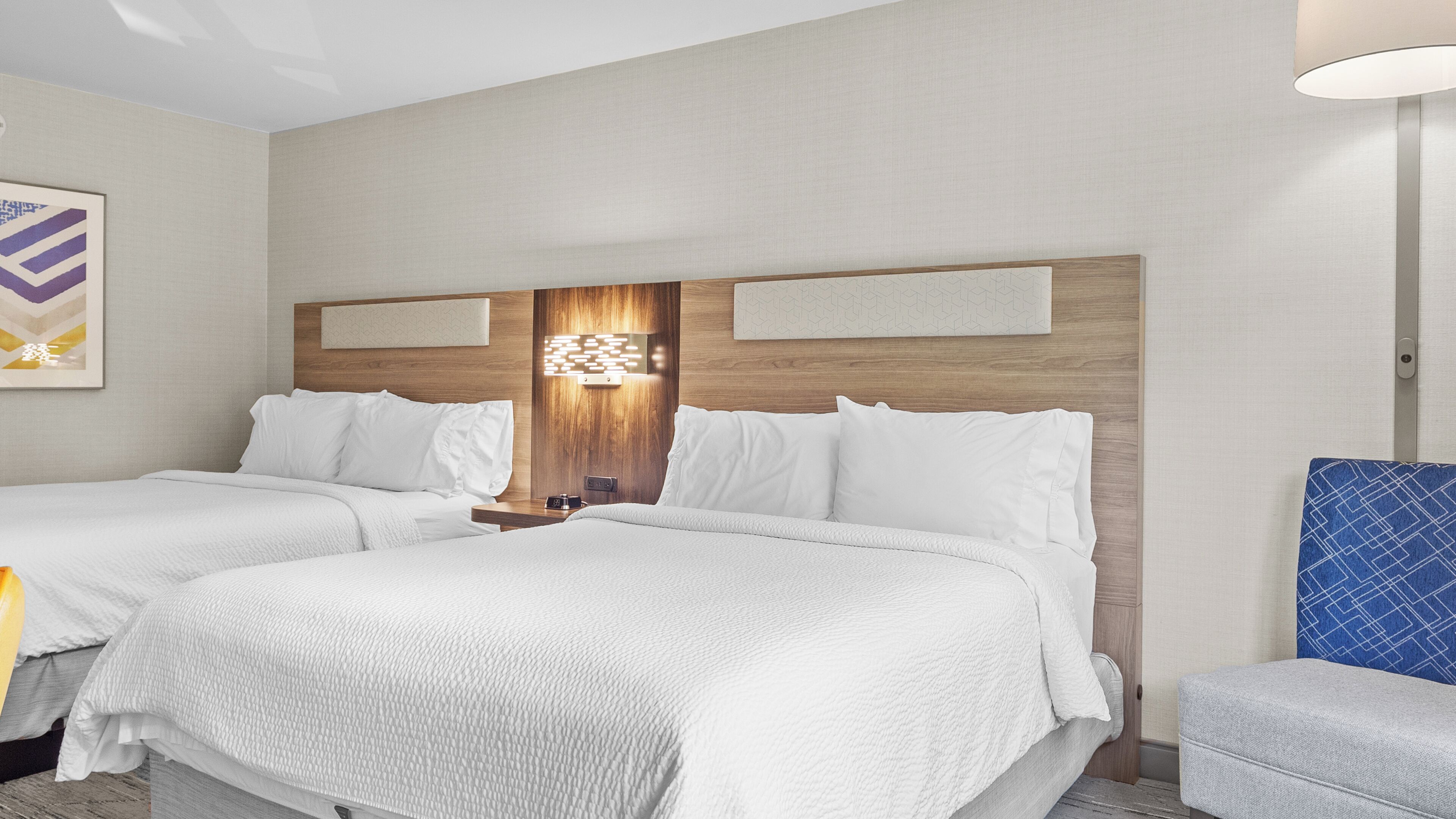A hotel room with two beds, a nightstand, a lamp, and a blue chair.