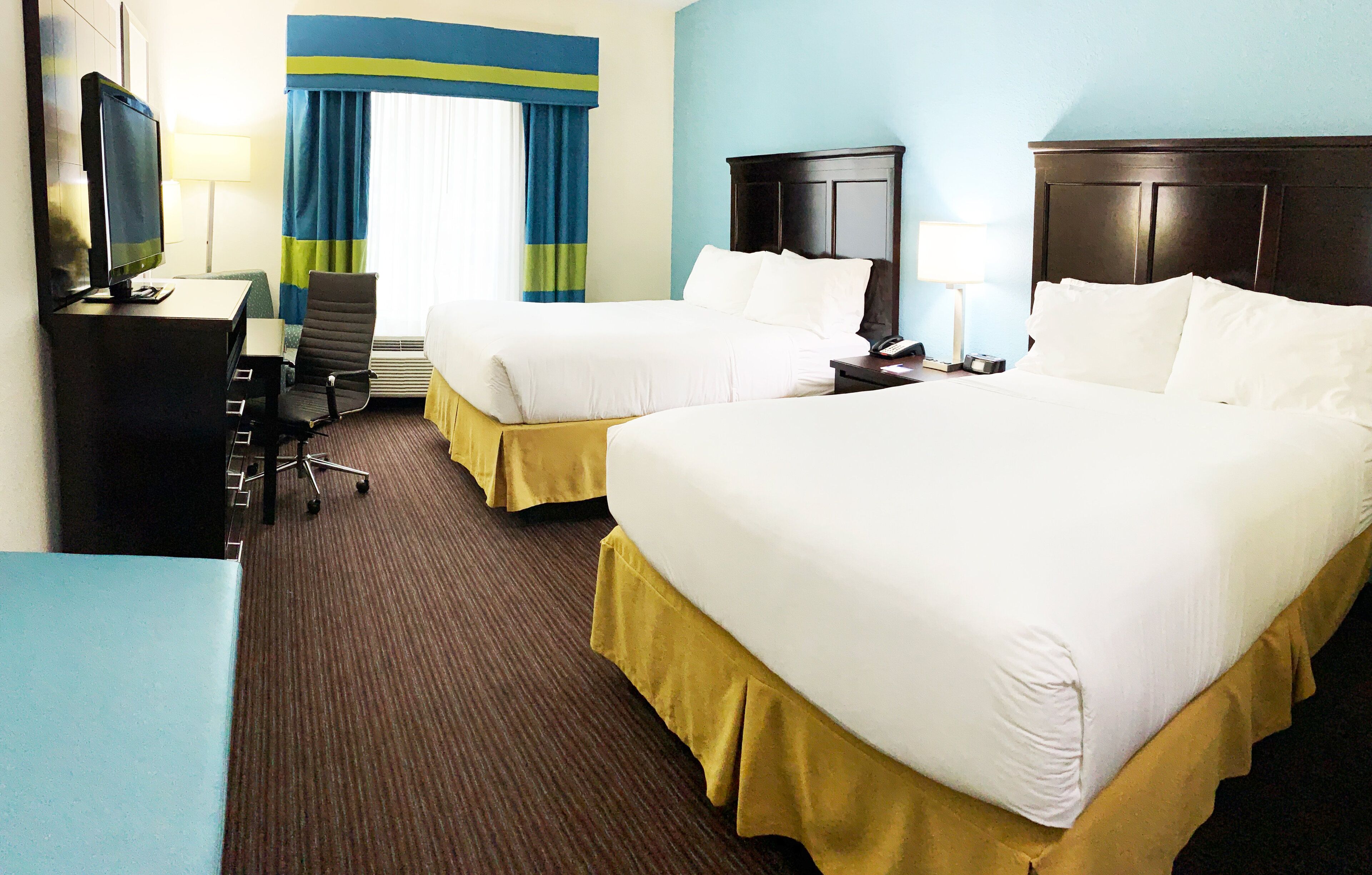 A hotel room with two beds, a desk with a computer, a chair, a nightstand with a lamp, and a window with curtains.
