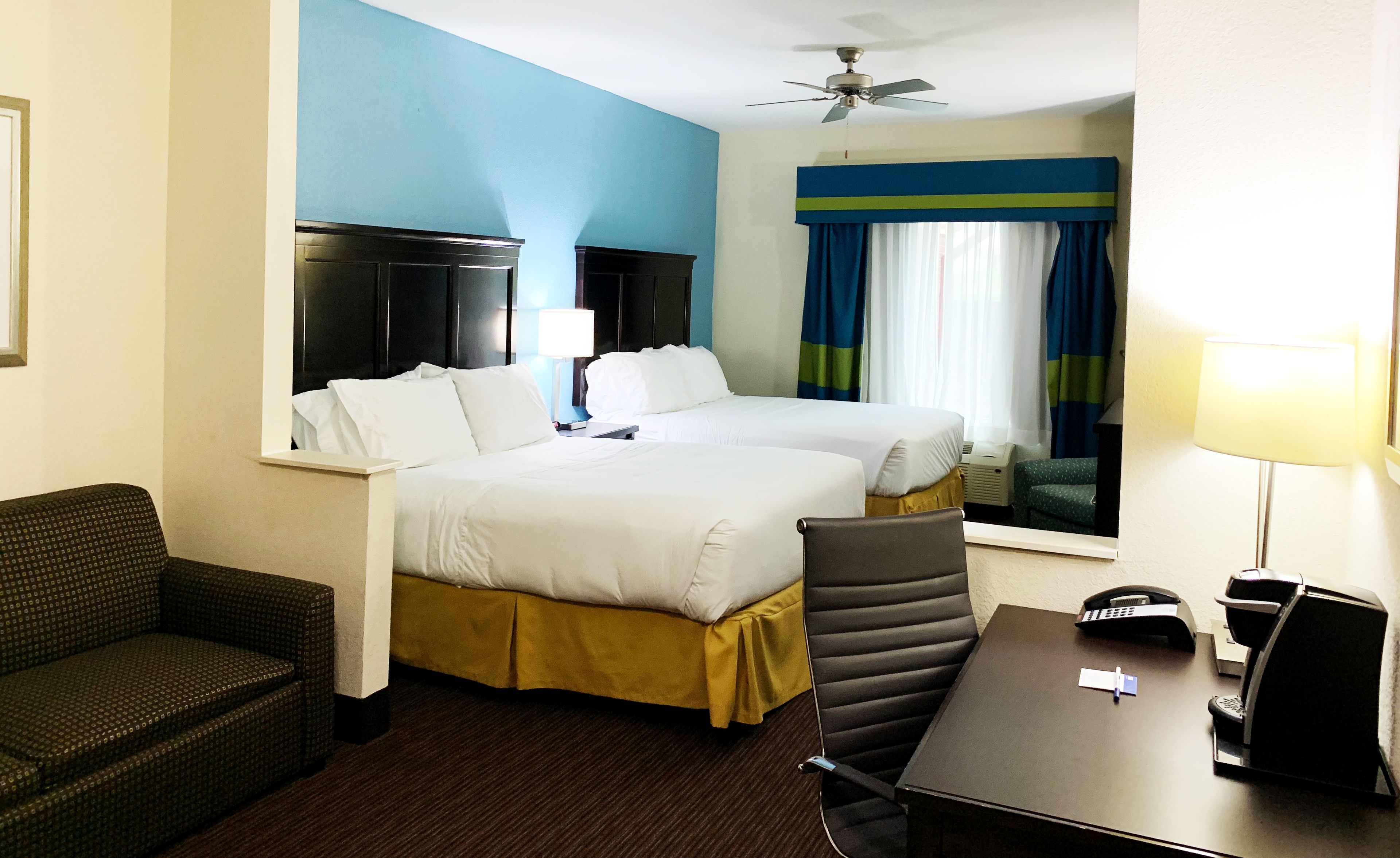 A hotel room with two beds, a desk, a chair, a desk lamp, a telephone, and a ceiling fan.