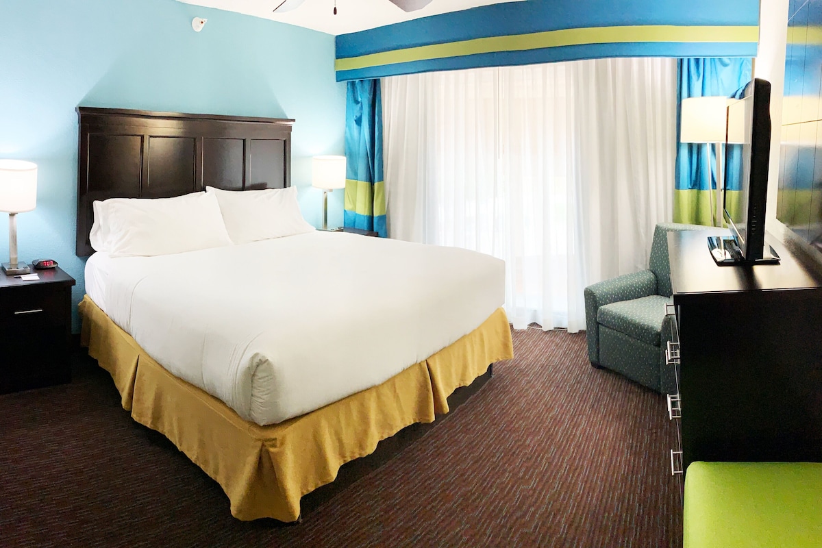 Ein Hotelzimmer mit einem großen Bett, Nachttischlampen, einem Schreibtisch mit Telefon und einem Fernseher auf einem Standfuß.
