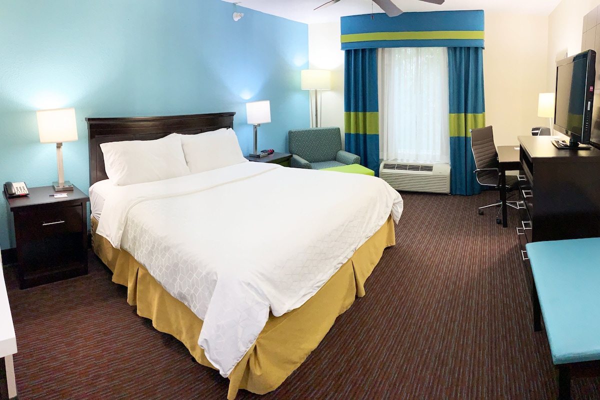 Ein Hotelzimmer mit einem großen Bett, Nachttischen, einem Schreibtisch, einem Sessel, einem Fernseher und einem Fenster mit Vorhängen.