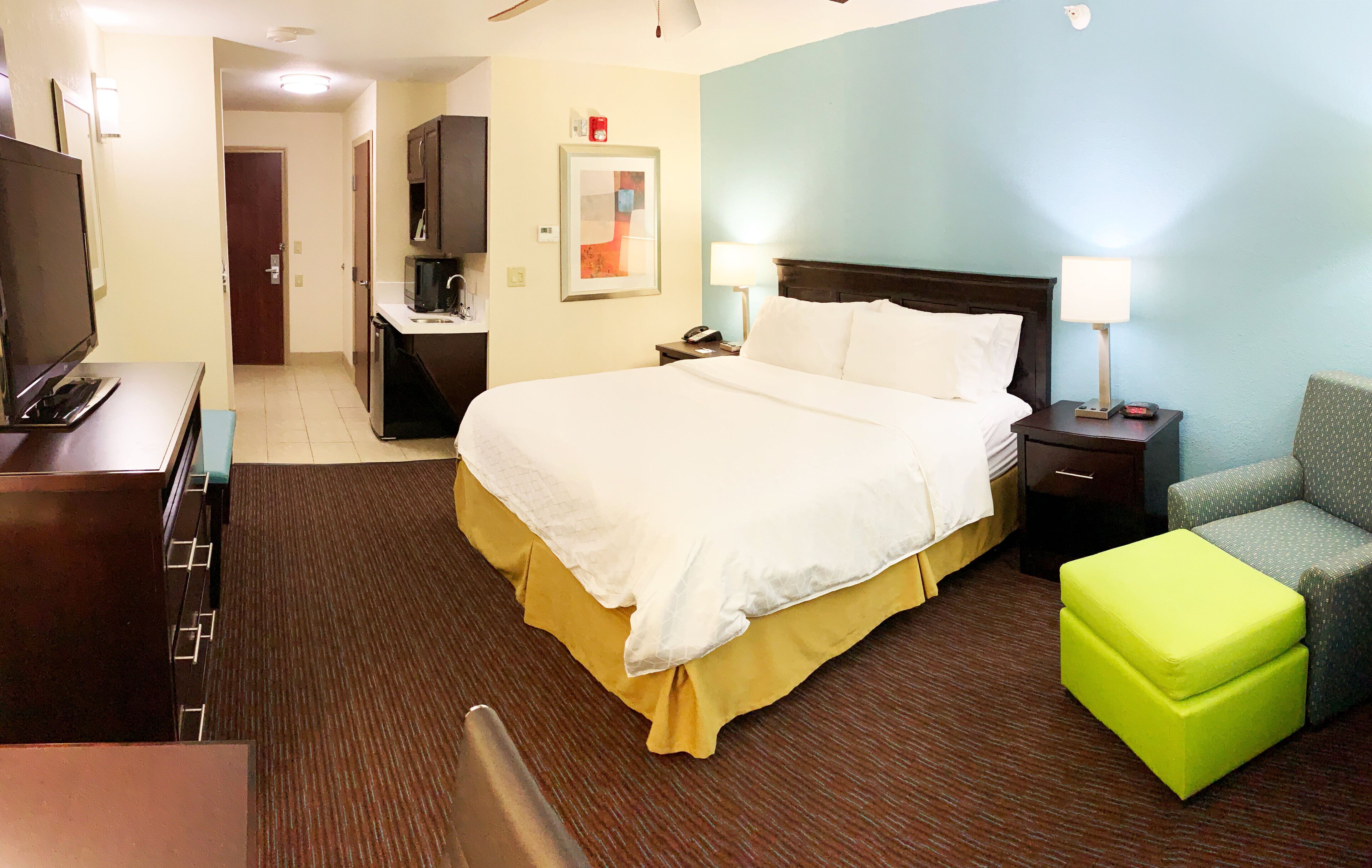 Quarto de hotel com cama, escrivaninha, cadeira, televisão e pequeno espaço de cozinha.