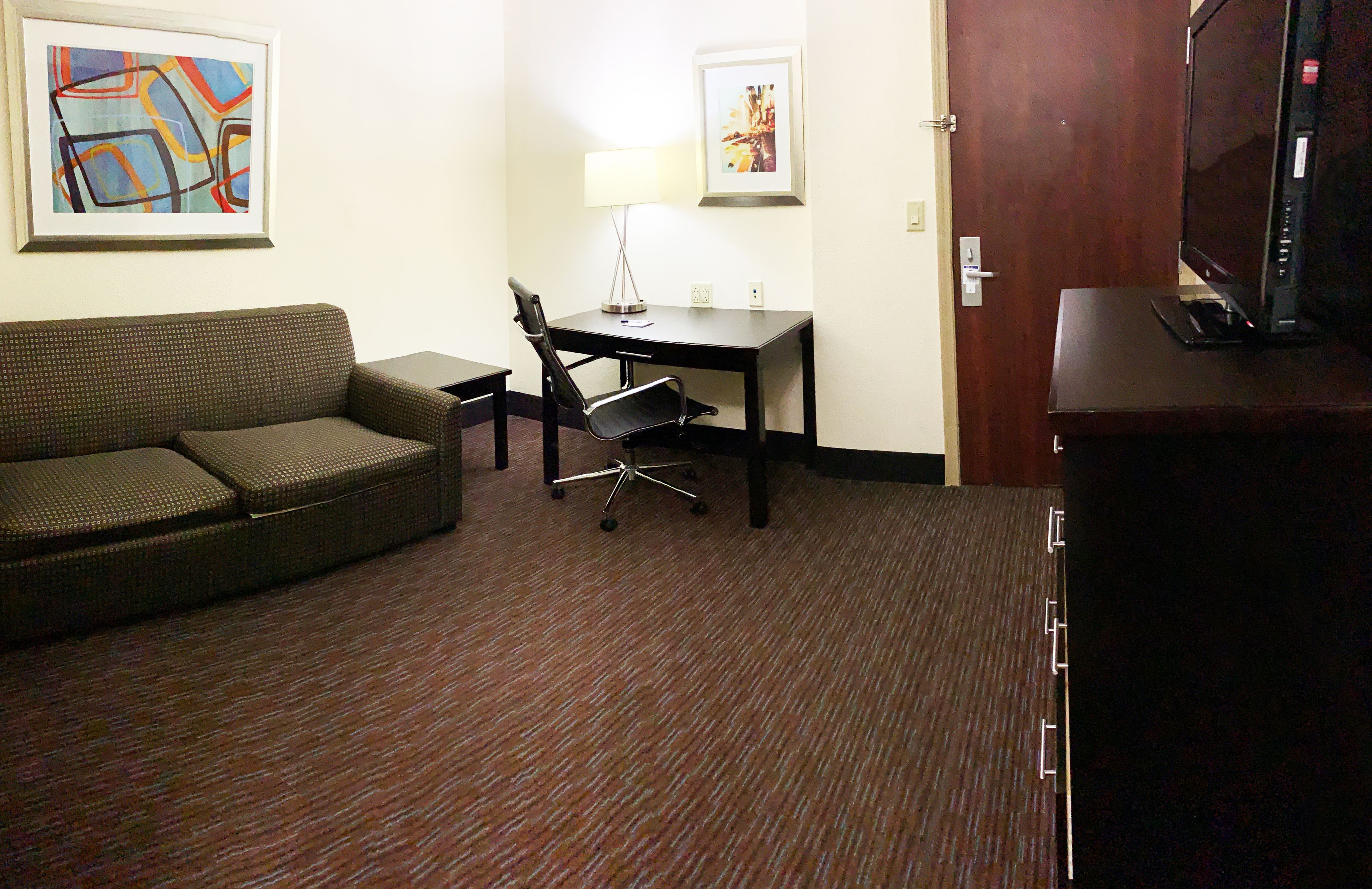 A hotel room with a sofa, a desk with a lamp, a television, and two framed pictures on the wall.