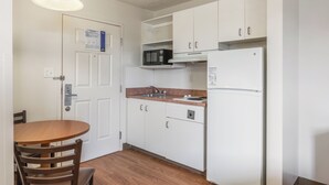 Ett kompakt kök med vita skåp, en mikrovågsugn, ett kylskåp och ett matbord i trä med stolar.