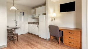 Een kleine keuken met witte kastjes, een magnetron, een koelkast, een eettafel met stoelen, een houten bureau met lades en een flatscreen televisie aan de muur.