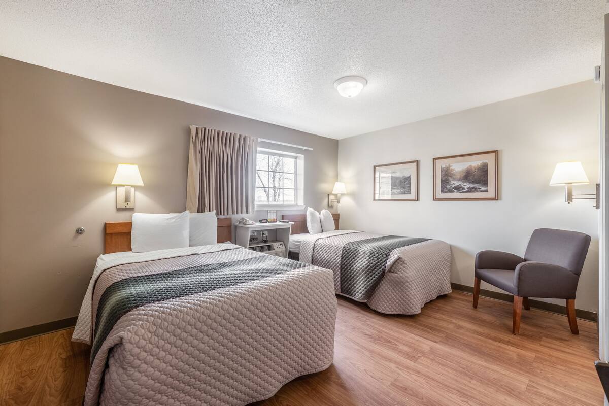 Ein Hotelzimmer mit zwei Betten, einem Schreibtisch, einem Stuhl und einem fensterbepflanzten Fenster.