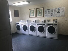 Un cuarto de lavandería con cinco lavadoras de carga frontal, un tablón informativo en la pared y piso de baldosas.