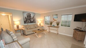 A living room with a beige sofa, a coffee table, and a TV. There are two windows and a door. - Cabana Beach Club by RVA (Longboat Key)