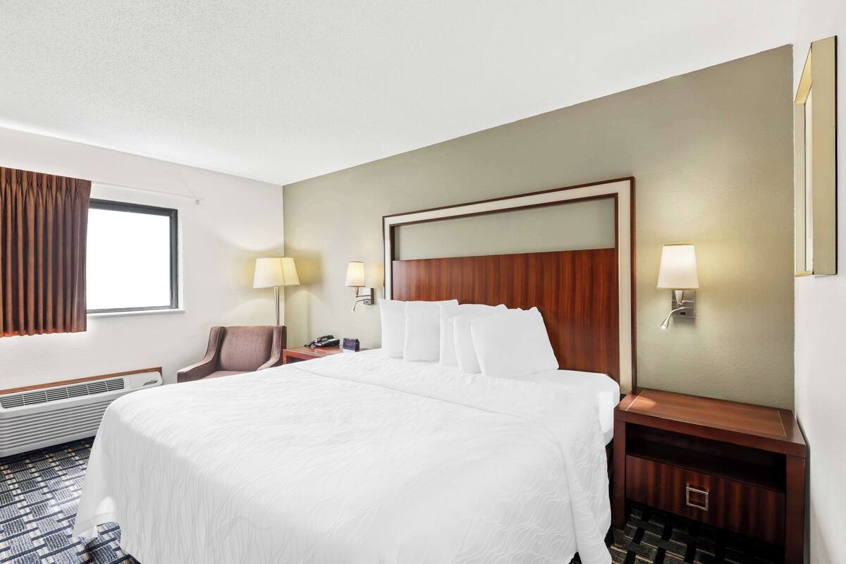 A hotel room with a large bed, two bedside tables with lamps, a chair, a desk with a phone, and a window with curtains.