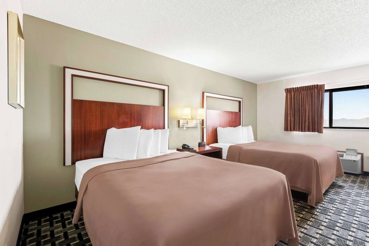 A hotel room with two beds, a nightstand with a lamp, a telephone, and a window with curtains.