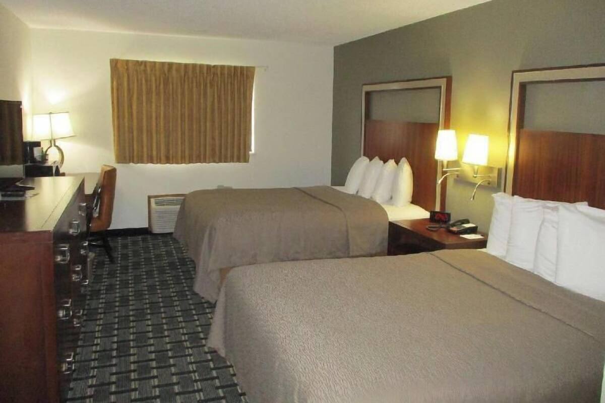 A hotel room with two beds, a desk with a lamp, a chair, a window with curtains, and a wall-mounted TV.