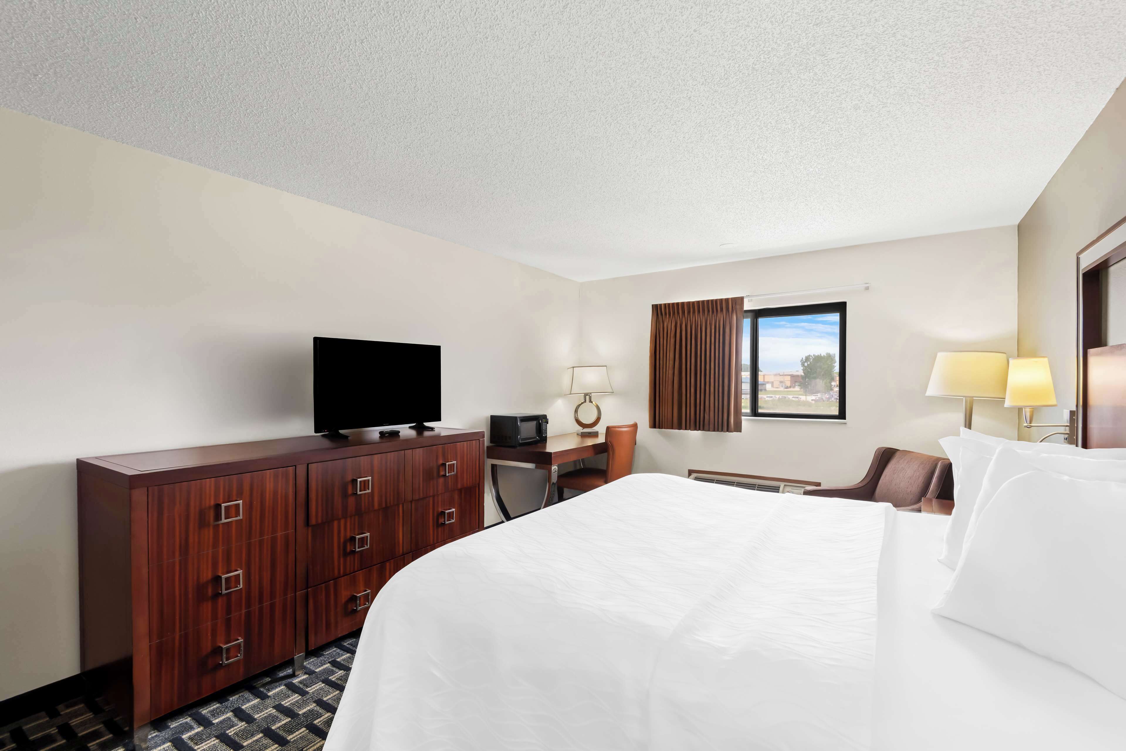 A hotel room with a large bed, a wooden dresser, a desk with a computer, a television, and a window with curtains.