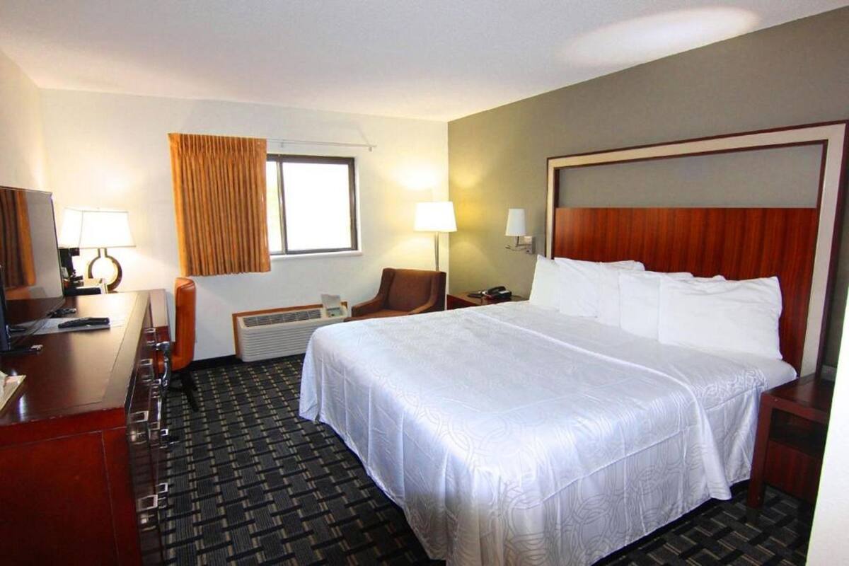 A hotel room with a large bed, a desk with a chair, a desk lamp, a chair, a window with curtains, and a wall-mounted heater.