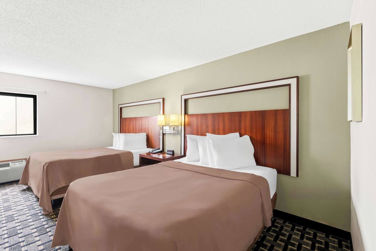 A hotel room with two beds, a nightstand with a lamp, and a window with blinds.