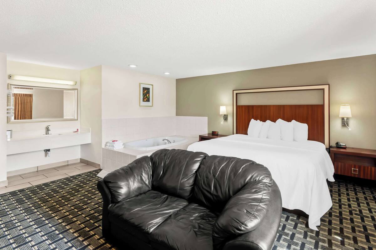 A hotel room with a bed, a black leather chair, a bathroom with a bathtub, and a large mirror.