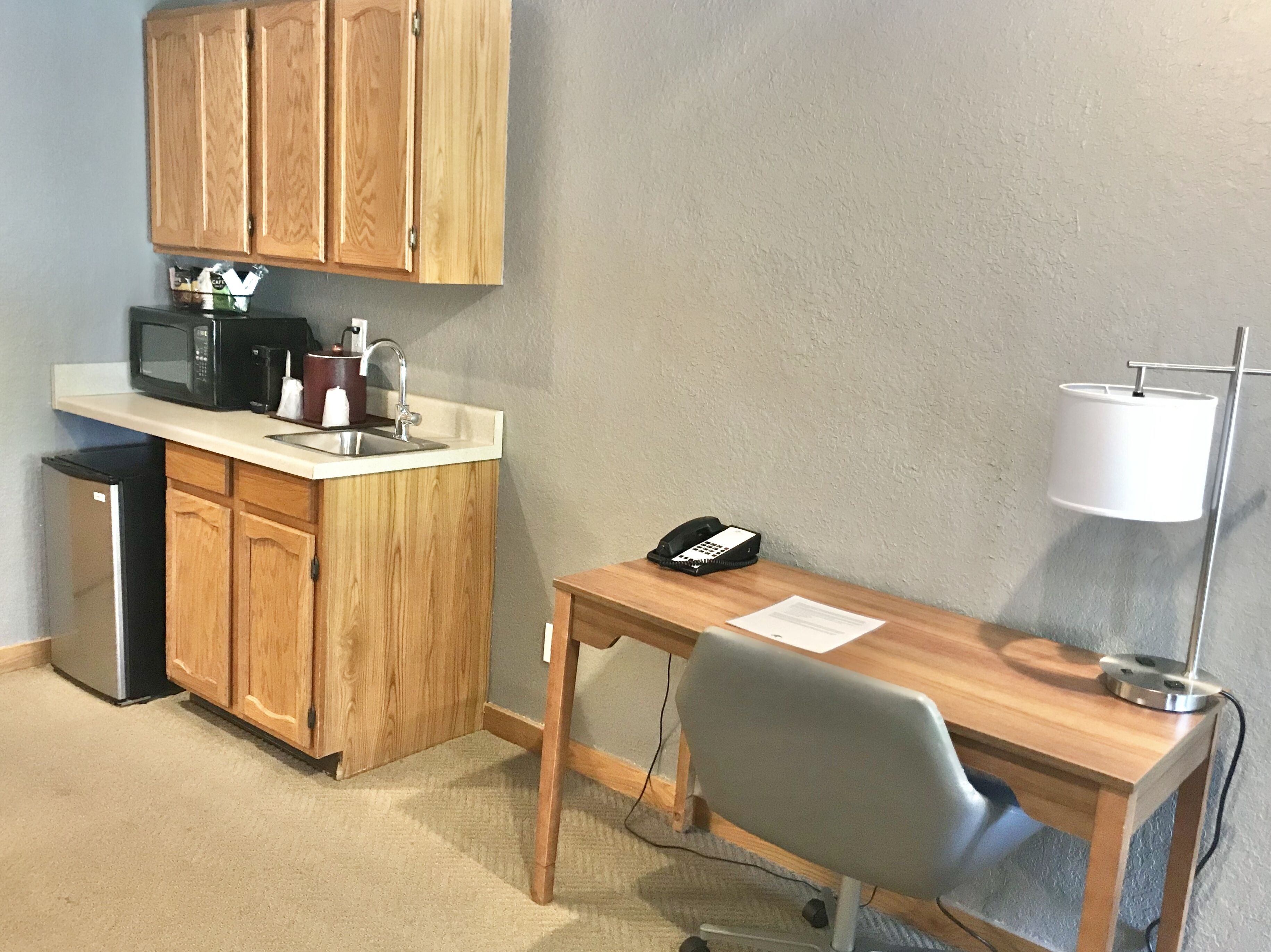 Una kitchenette compacta con microondas, cafetera y lavaplatos. Hay un escritorio con teléfono y lámpara, además de un refrigerador pequeño.
