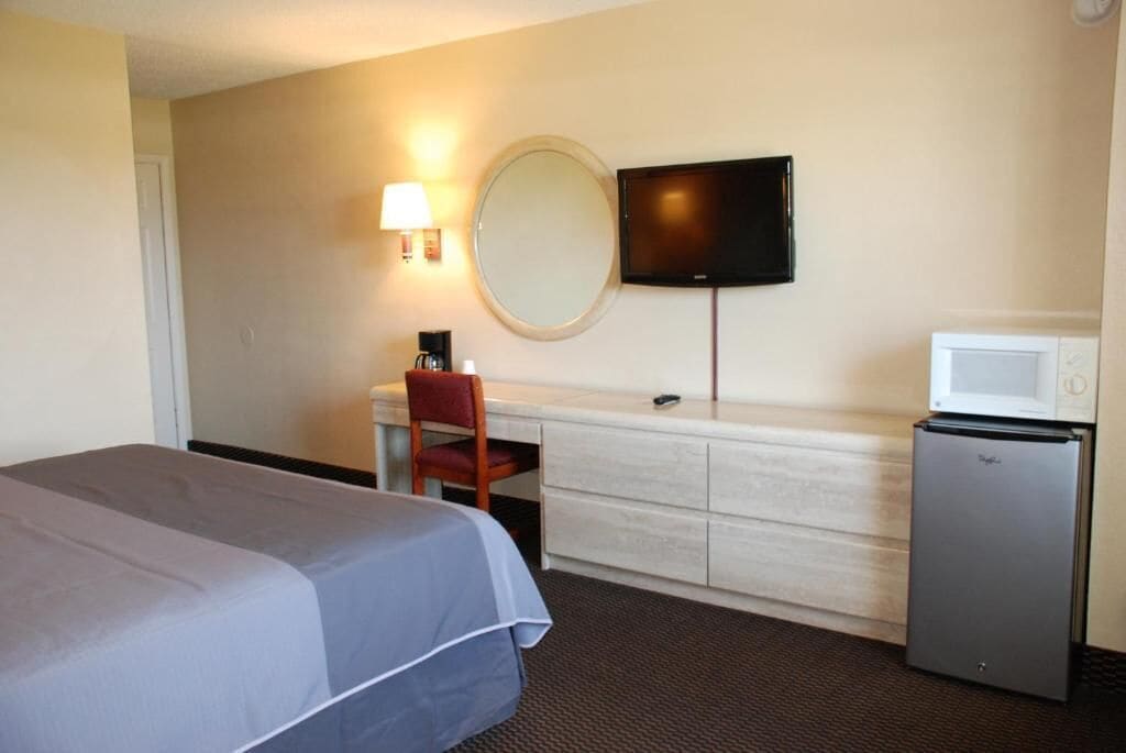 A hotel room with a bed, a desk with a chair, a television, a mirror, and a refrigerator.