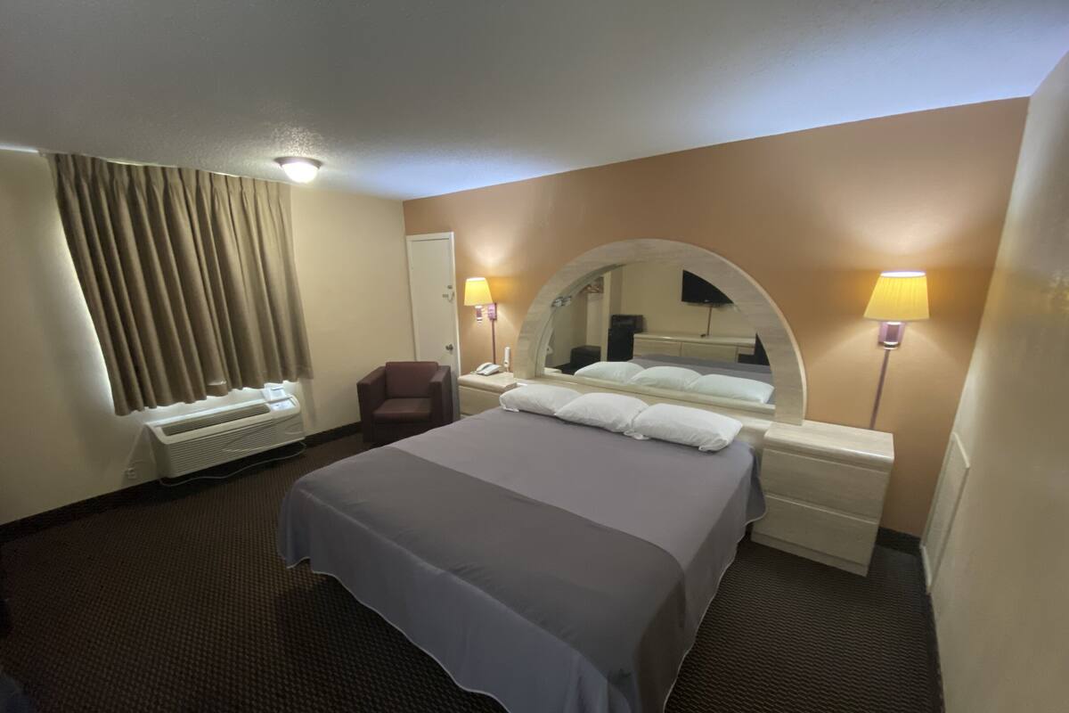 A hotel room with a bed, a chair, a desk with a lamp, and a mirror.