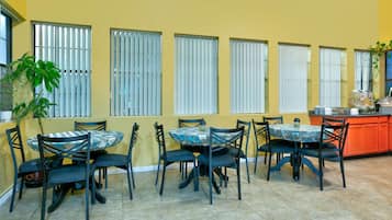 A dining area with four round tables, each surrounded by four chairs. The walls are yellow, and there are six windows with blinds. A potted plant is visible on the left side.