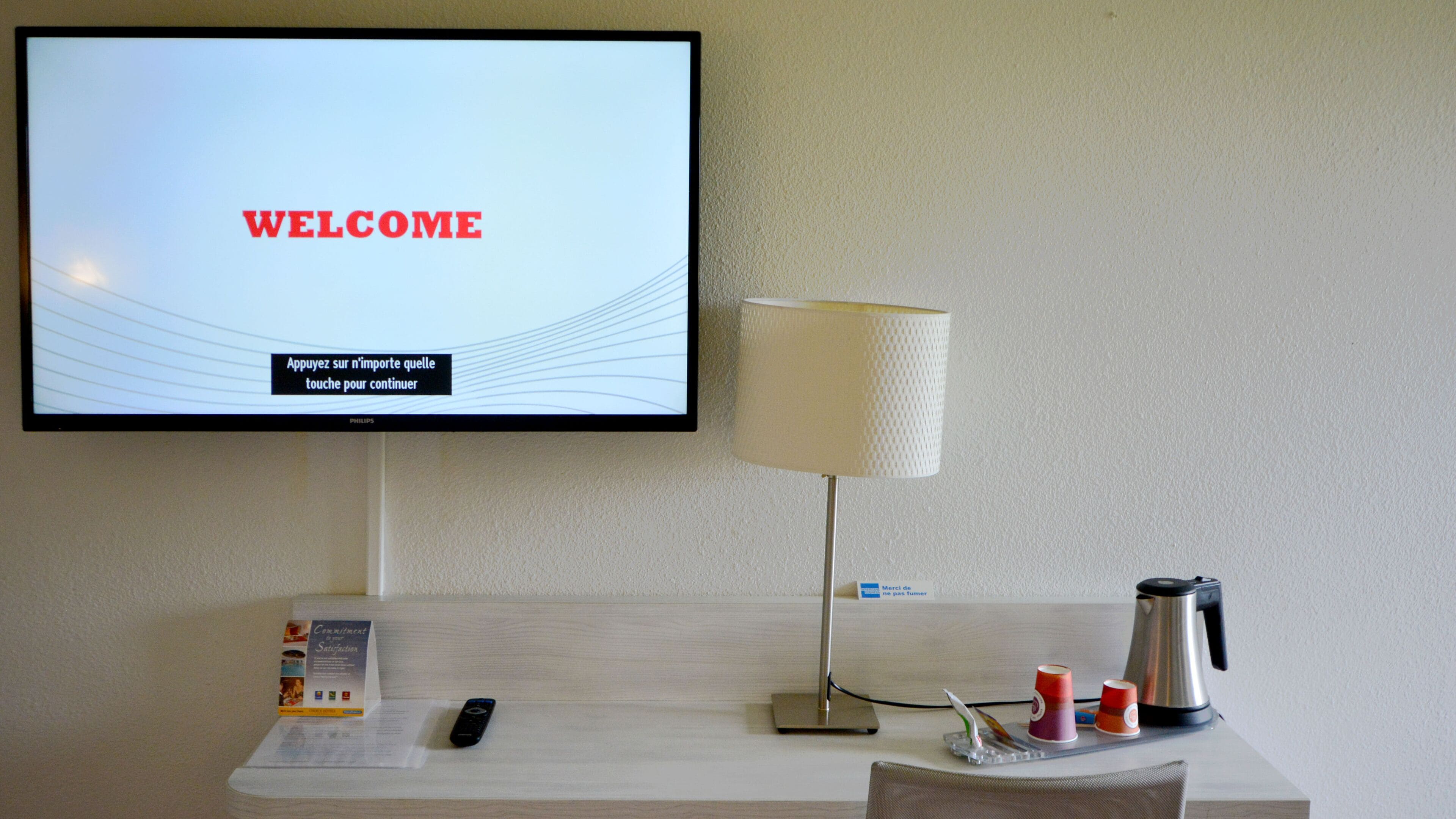 A hotel room with a television displaying a welcome message, a lamp, a remote control, a coffee maker, and a small table with a menu and a remote control.