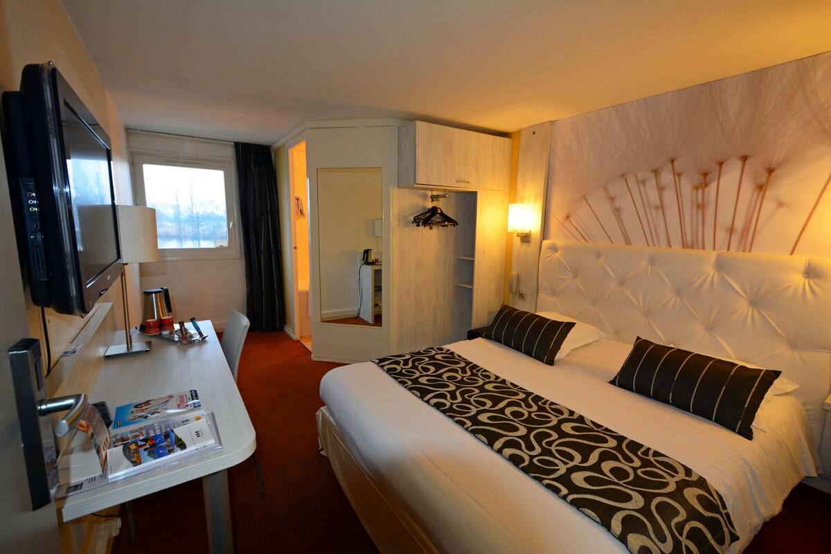 Ett hotellrum med en säng, ett skrivbord med en platt-TV, en spegel och ett fönster.