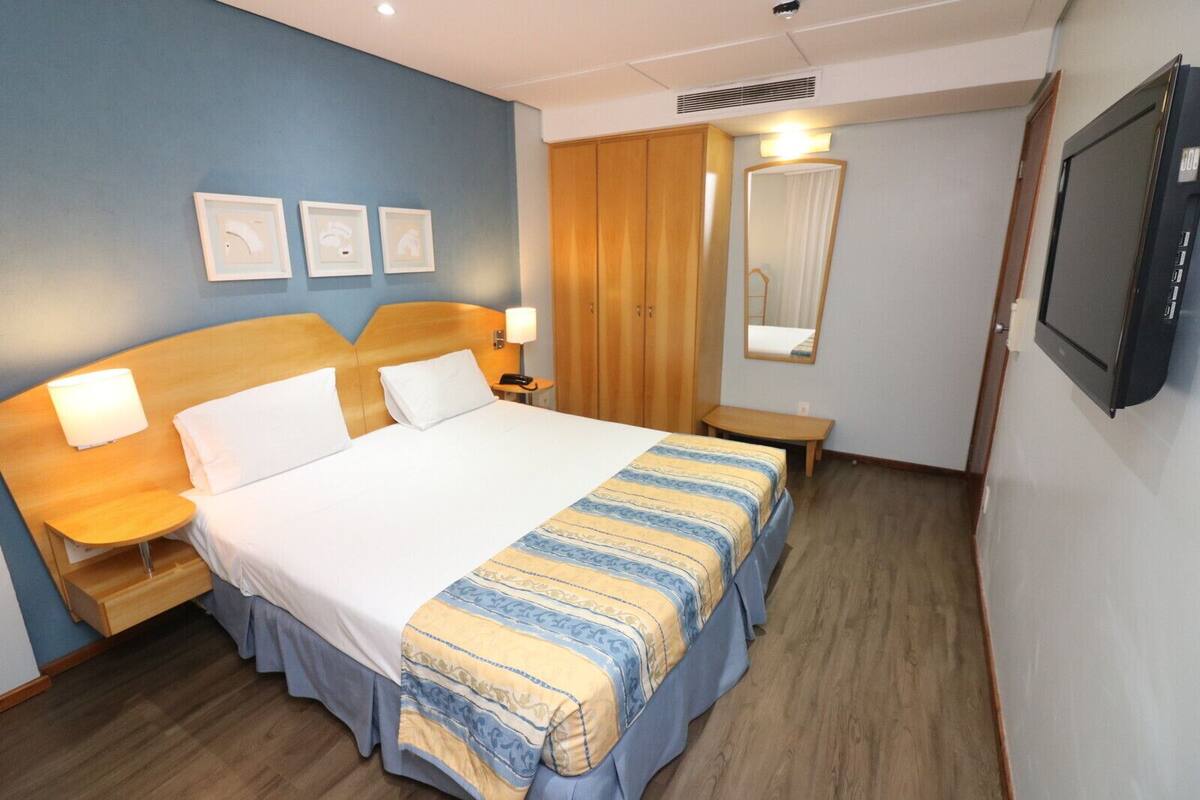 Ein Hotelzimmer mit einem großen Bett, Nachttischen inklusive Lampen, einem Holzschrank, einem Spiegel und einem Fernseher.
