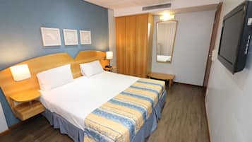 A hotel room with a large bed, bedside tables with lamps, a wooden wardrobe, a mirror, and a television.