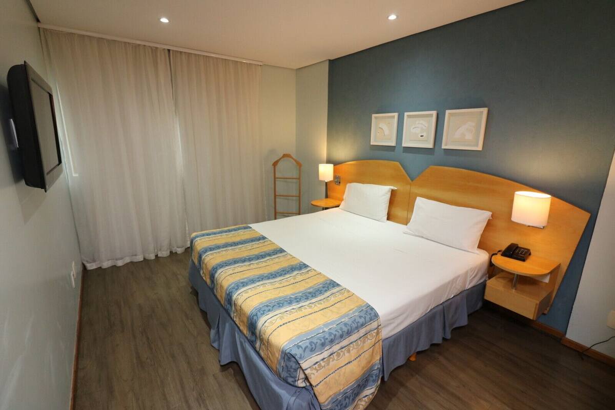 Ein Hotelzimmer mit einem großen Bett, einem Nachttisch mit Lampe, einem Telefon und einem an der Wand befestigten Fernseher.