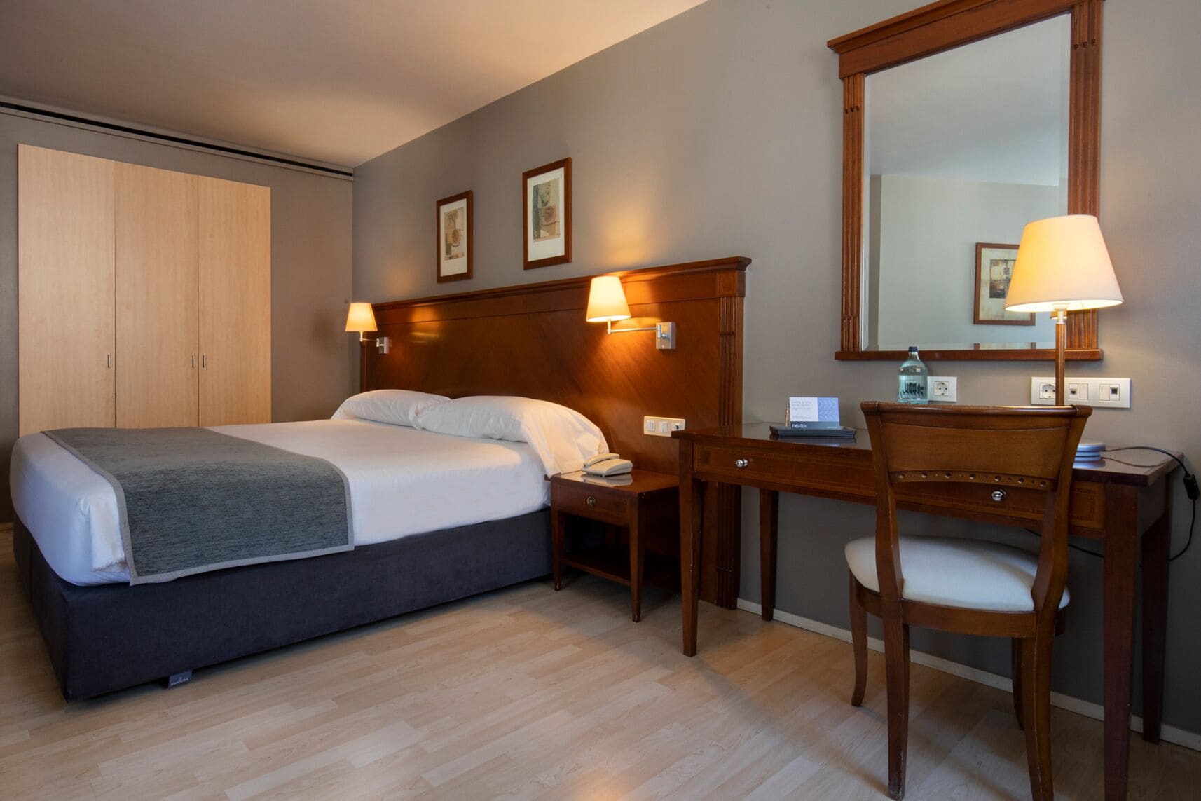 A hotel room with a bed, bedside table, desk, chair, and a large mirror.