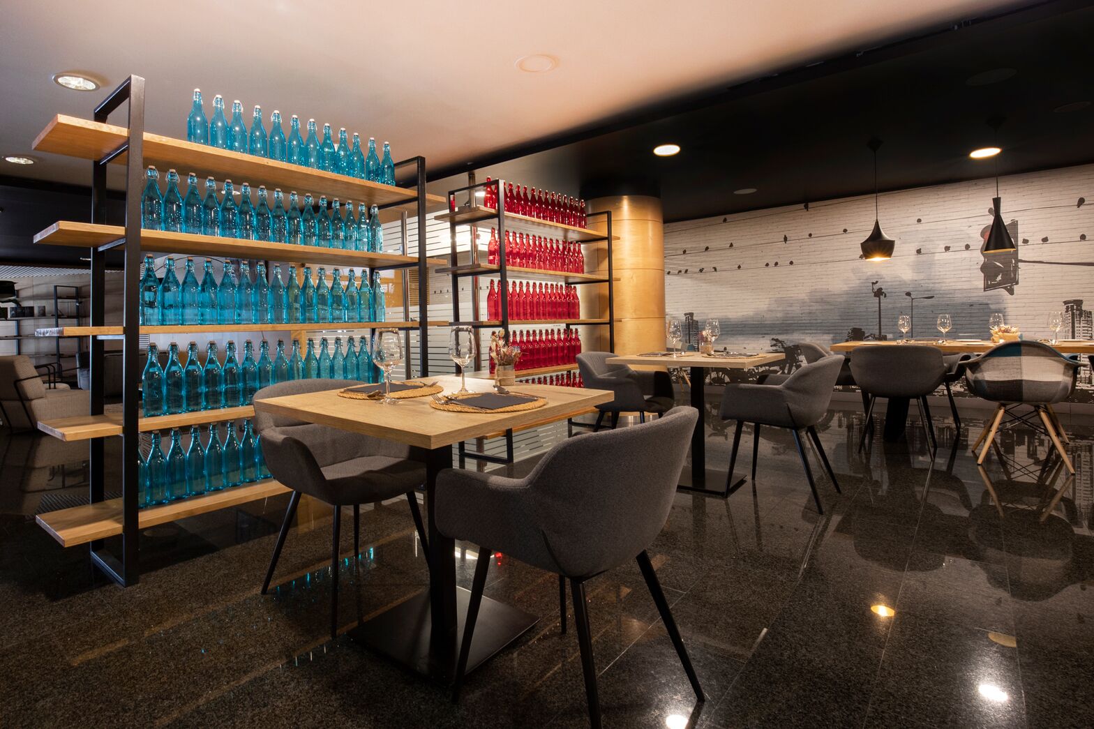 A modern restaurant interior with wooden tables and chairs, shelves with blue and red bottles, and a wall with hanging lights.