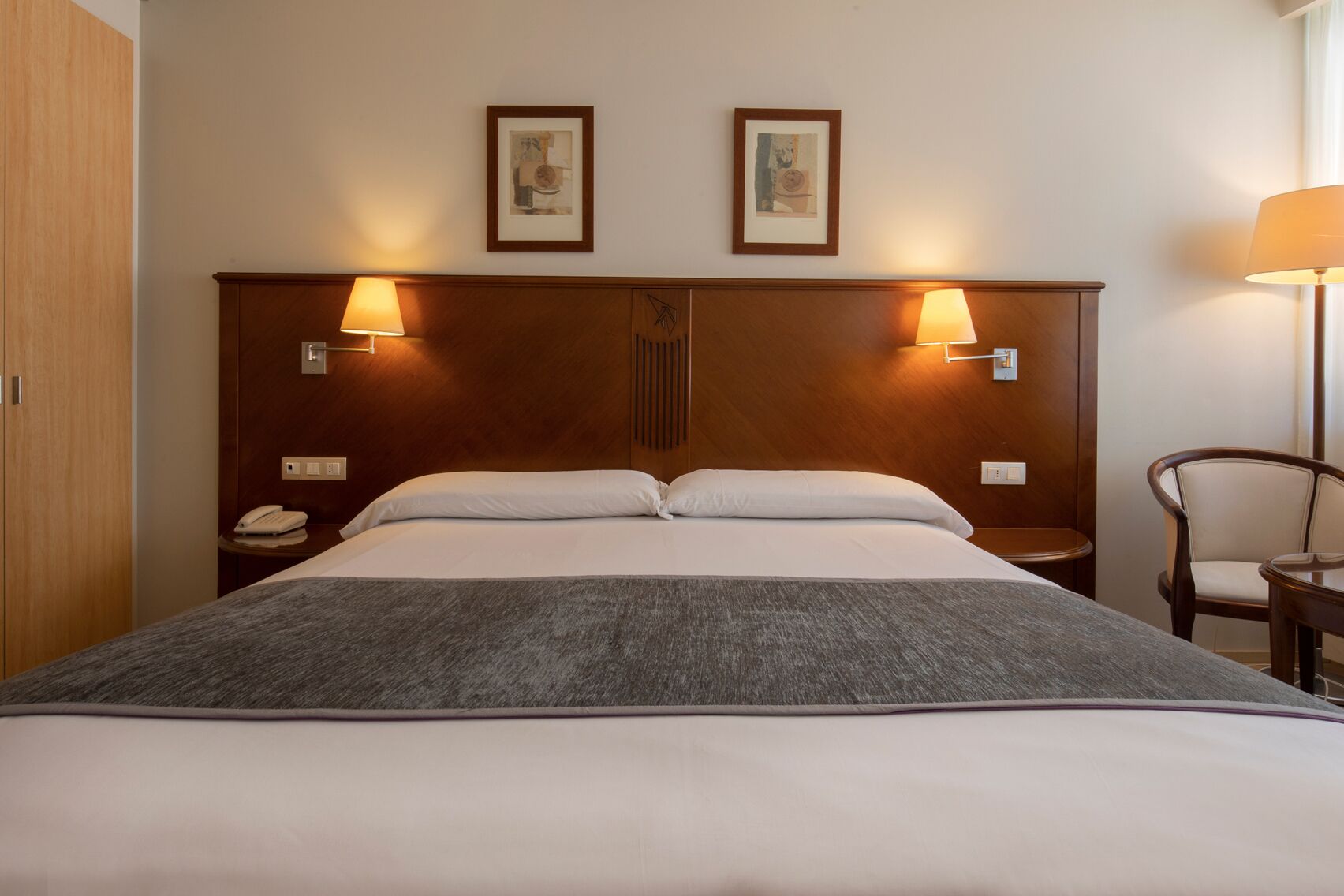A hotel room with a bed, two bedside lamps, a nightstand with a phone, and a chair.