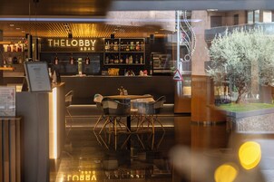 Un espace bar agrémenté d’un panneau néon indiquant « THE LOBBY & BAR », de diverses bouteilles exposées sur des étagères, et d’un coin salon avec une table ronde et des chaises.