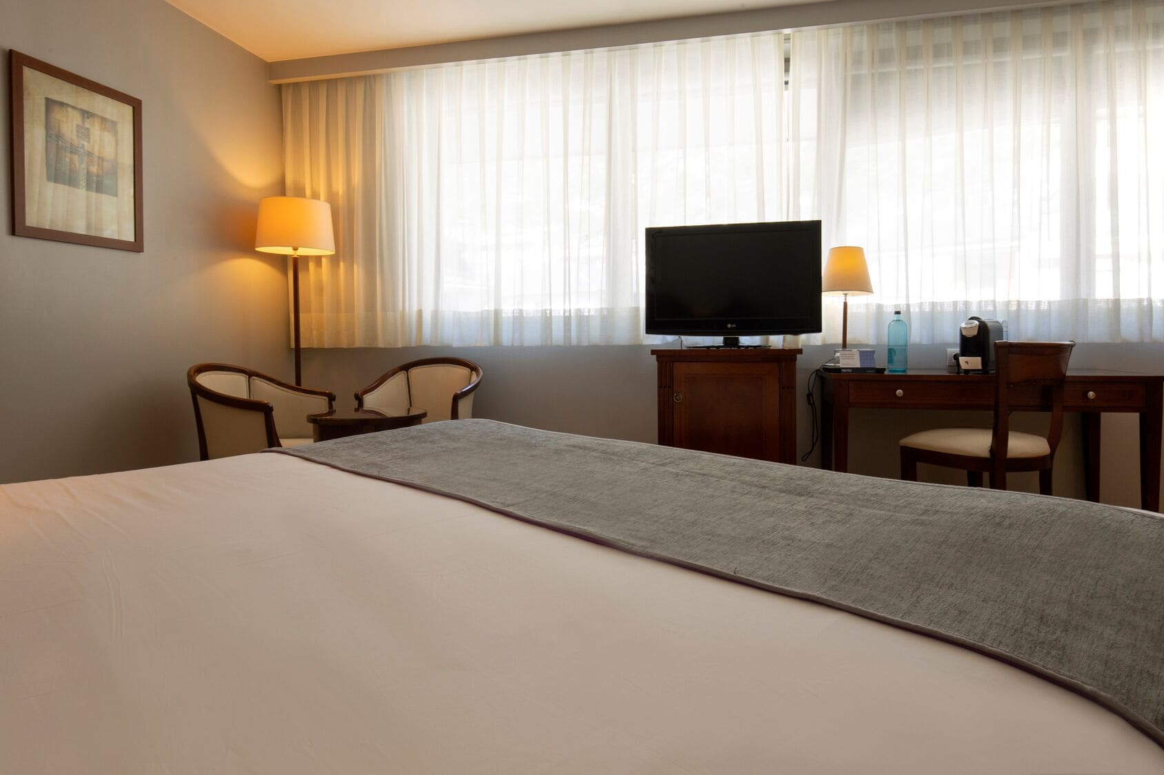 A hotel room with a bed, a desk, two chairs, a television, and a lamp.