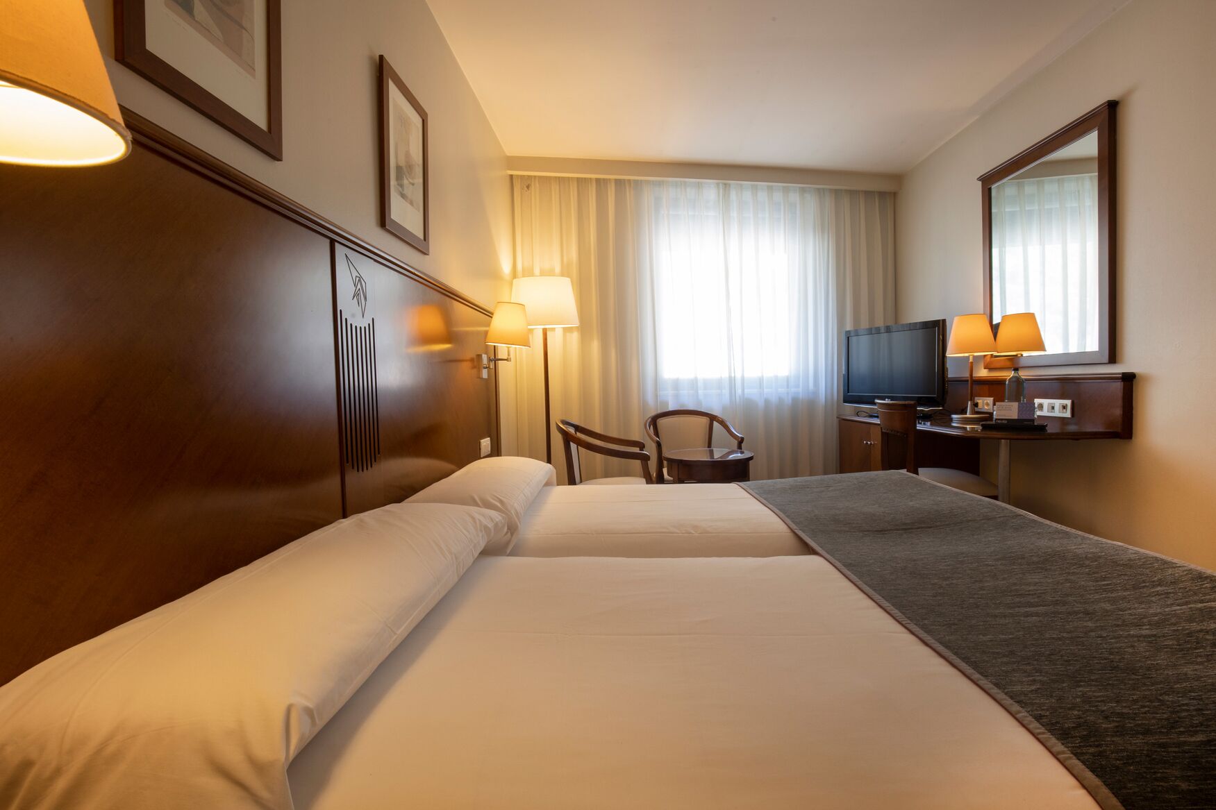 Habitación de hotel con dos camas, un escritorio con televisor, una silla y un ventanal con cortinas.