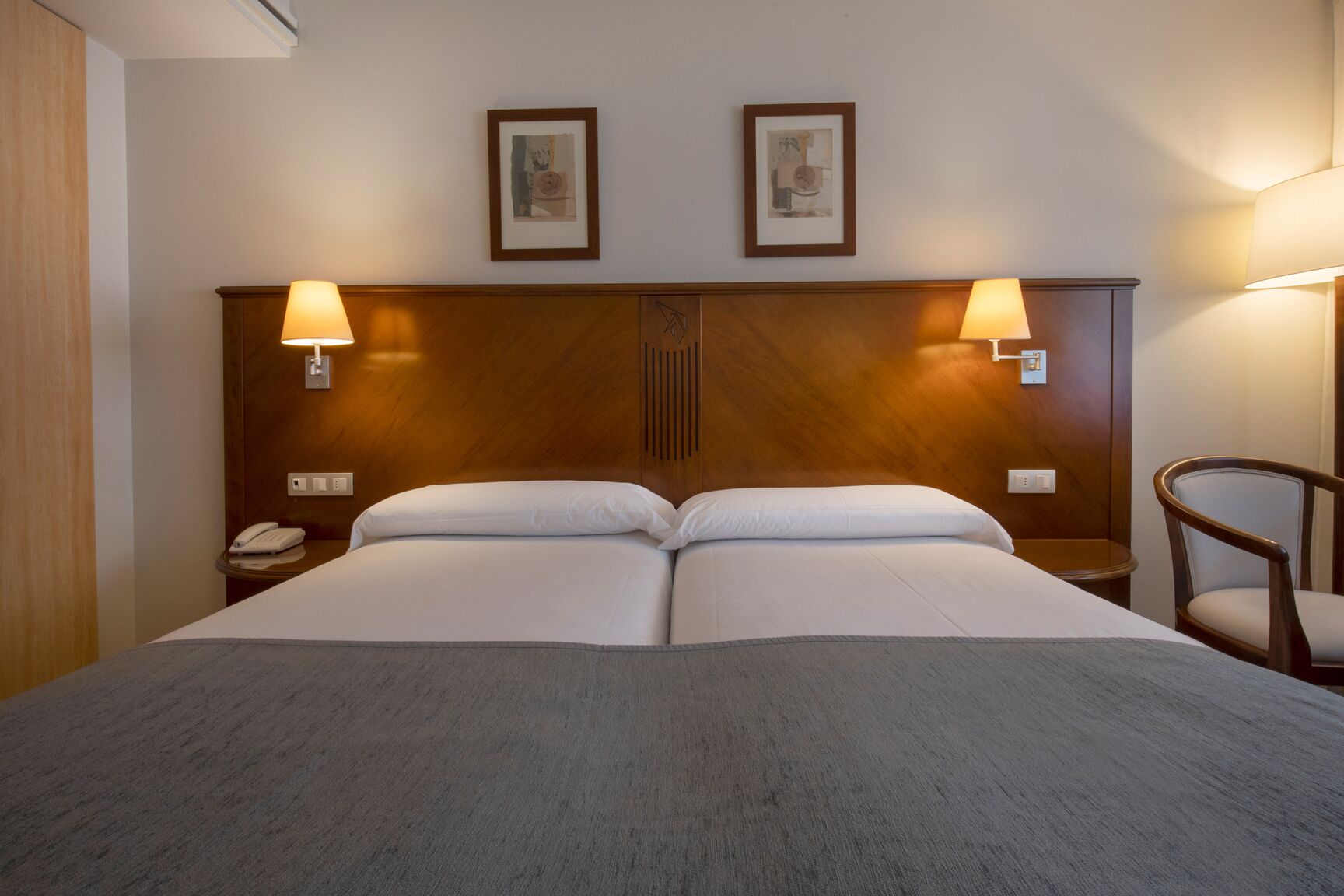Una cama doble con cabecera de madera, dos lámparas de noche, una mesita de noche con un teléfono y una silla.