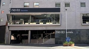 Um edifício moderno de hotel com fachada de vidro, entrada visível e uma placa com a inscrição 'nexta Escaldes'.