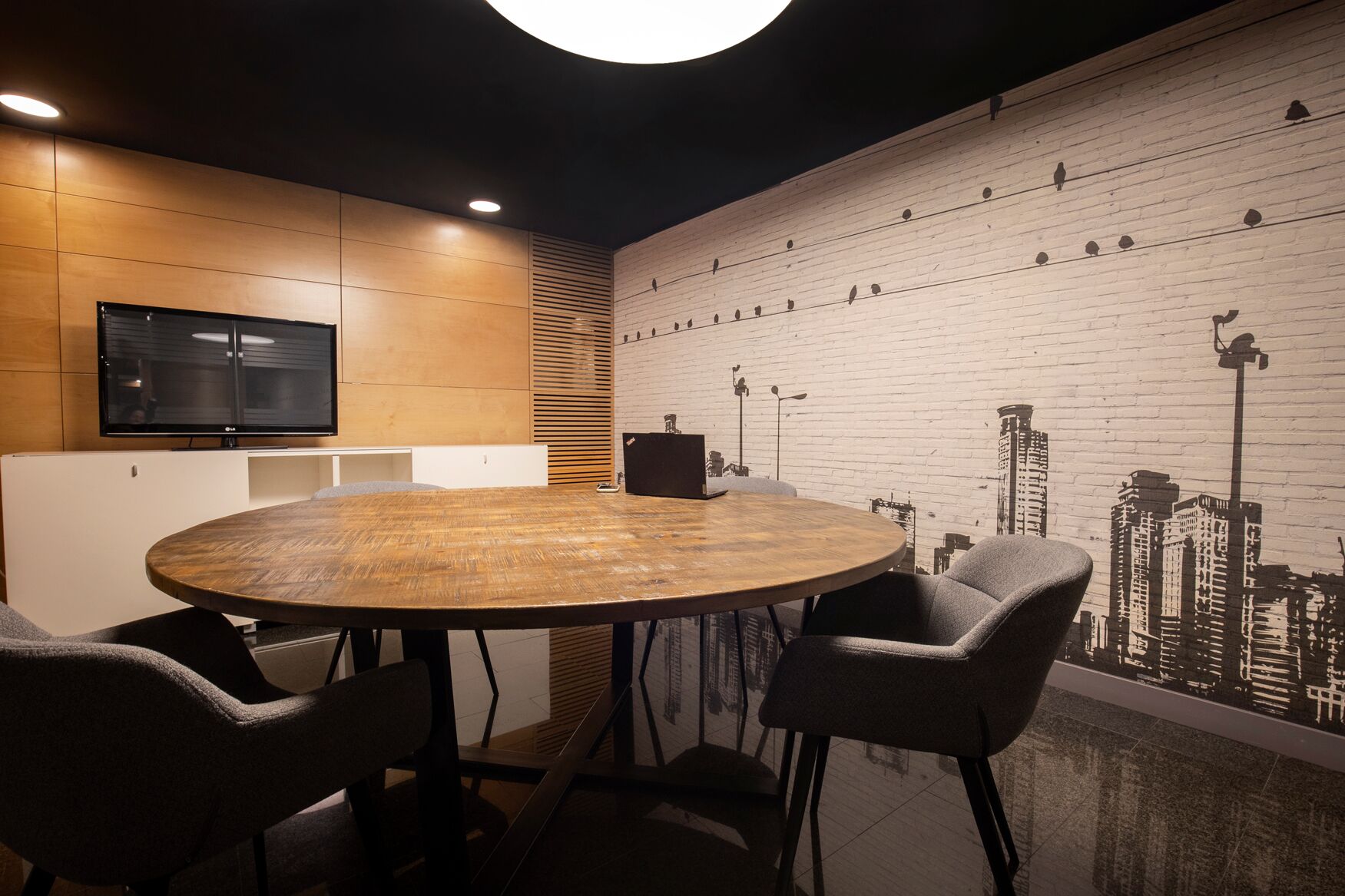 A conference room with a round wooden table, four chairs, a TV, and a cityscape mural on the wall.