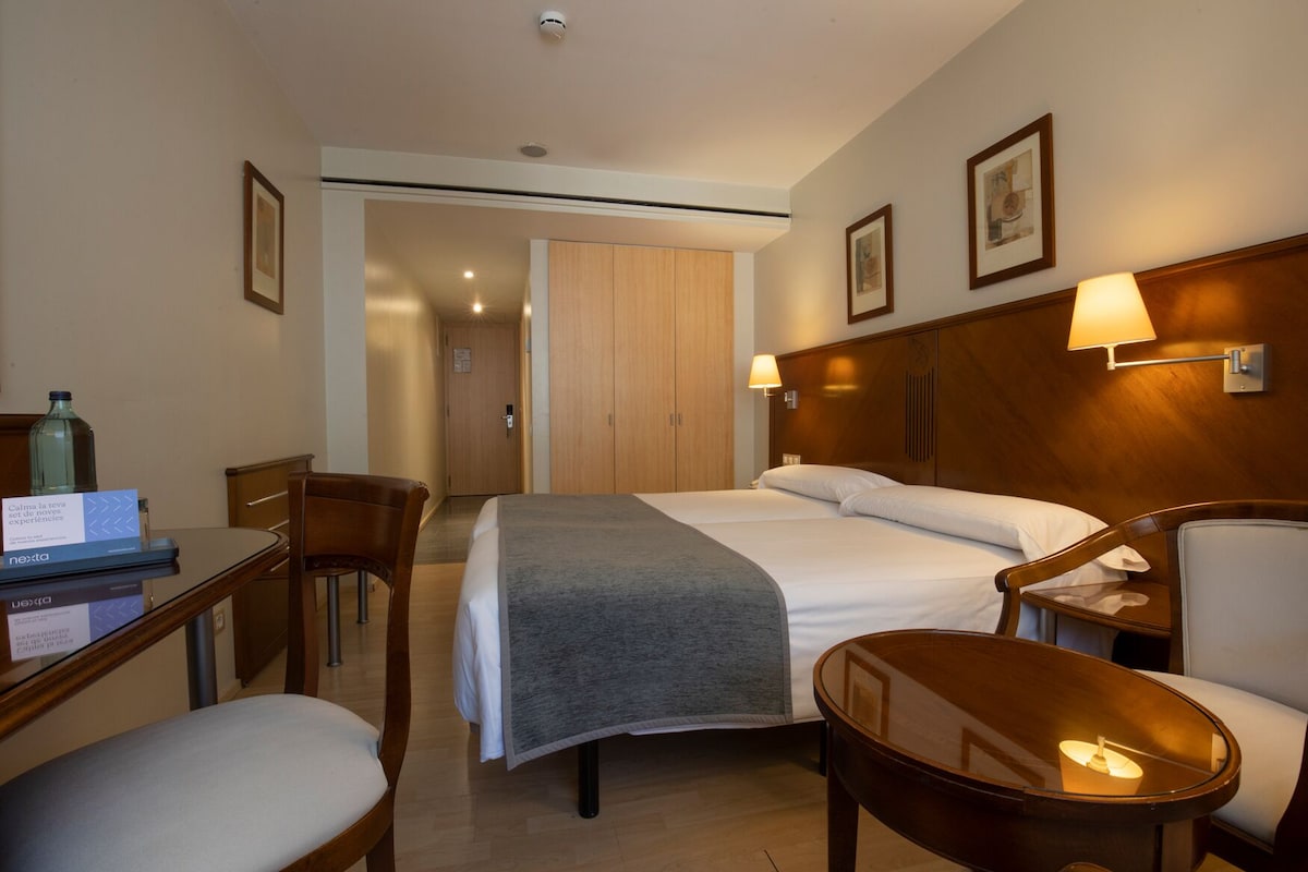 Een hotelkamer met een bed, nachtkastjes, een bureau, een stoel en een deur naar een gang.