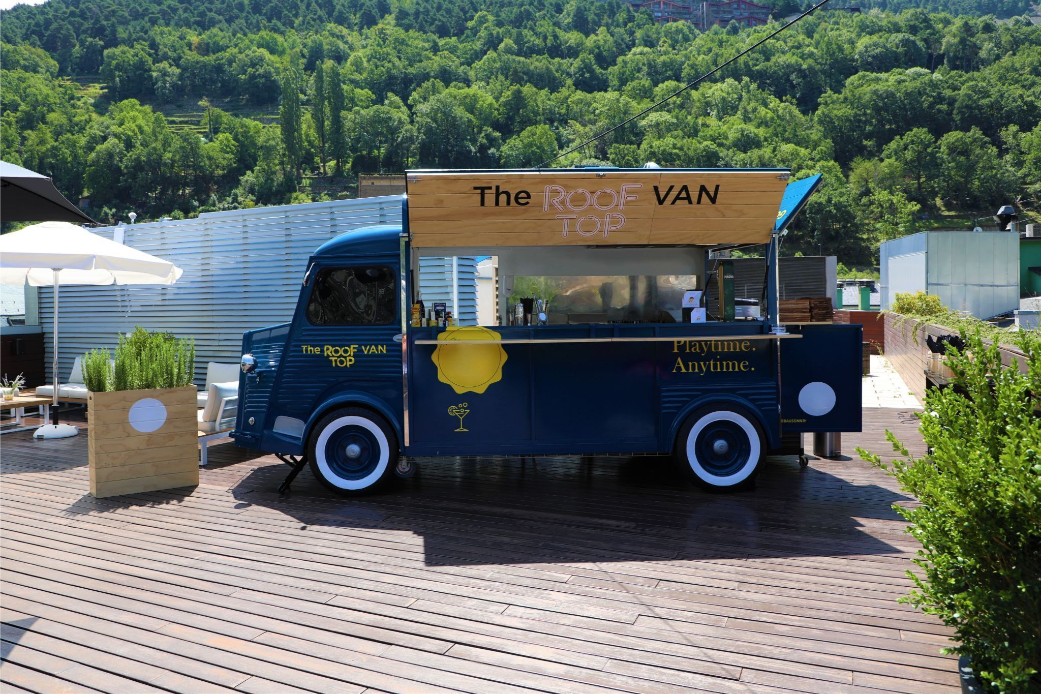 A food truck named "The Roof Van Top" parked on a wooden deck with a view of a green hillside in the background.