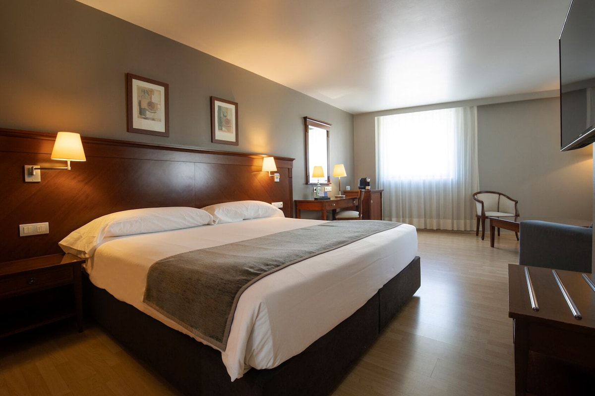 Een hotelkamer met een groot bed, bedlampjes, een bureau met stoel, een televisie en foto's in lijsten aan de muur.