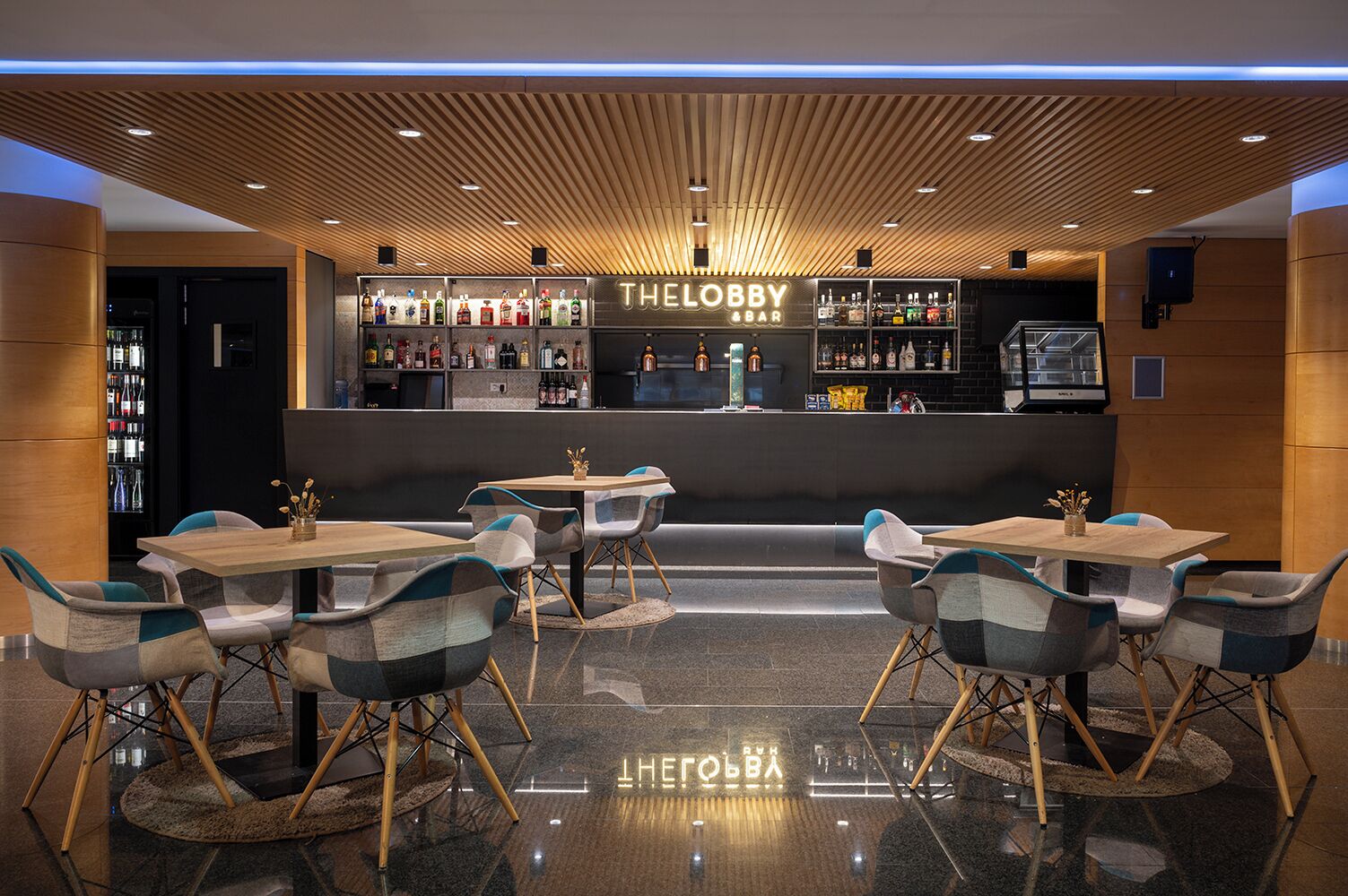 Zona bar dal design moderno, con soffitto in legno e diverse bottiglie di liquori sugli scaffali. Sono disposti tavoli e sedie per i clienti.