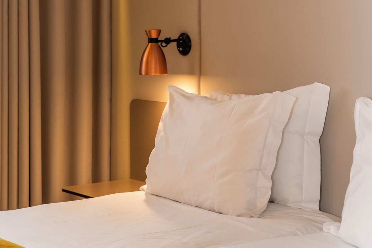 A neatly made bed with white linens and a yellow throw blanket, a wall-mounted lamp, and a curtain on the left.